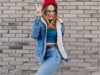 Ecstatic blonde girl in red hat standing in confident pose beside brick wall. Outdoor shot of cute caucasian woman in jeans and denim jacket having fun on the street.