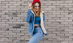 Ecstatic blonde girl in red hat standing in confident pose beside brick wall. Outdoor shot of cute caucasian woman in jeans and denim jacket having fun on the street.