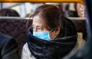 Older Asian woman wearing black fur scarf and surgical pollution mask sitting on a bu
